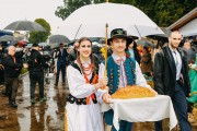 Kobieta oraz mężczyzna w strojach ludowych niosą chleb dożynkowy