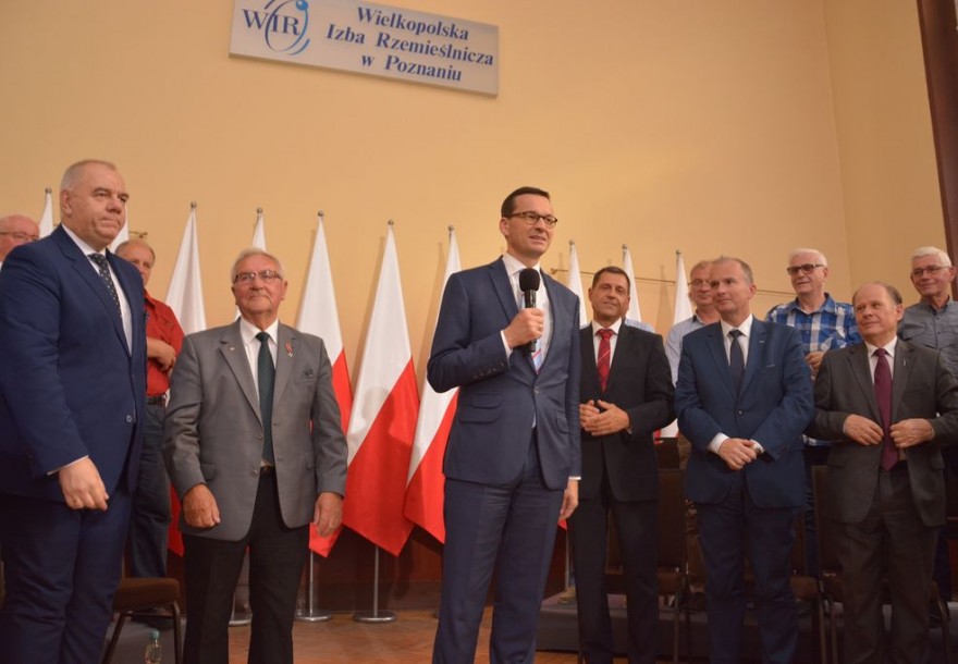 Premier Mateusz Morawiecki w otoczeniu przedsiębiorców, za plecami premiera biało-czerwone flagi.