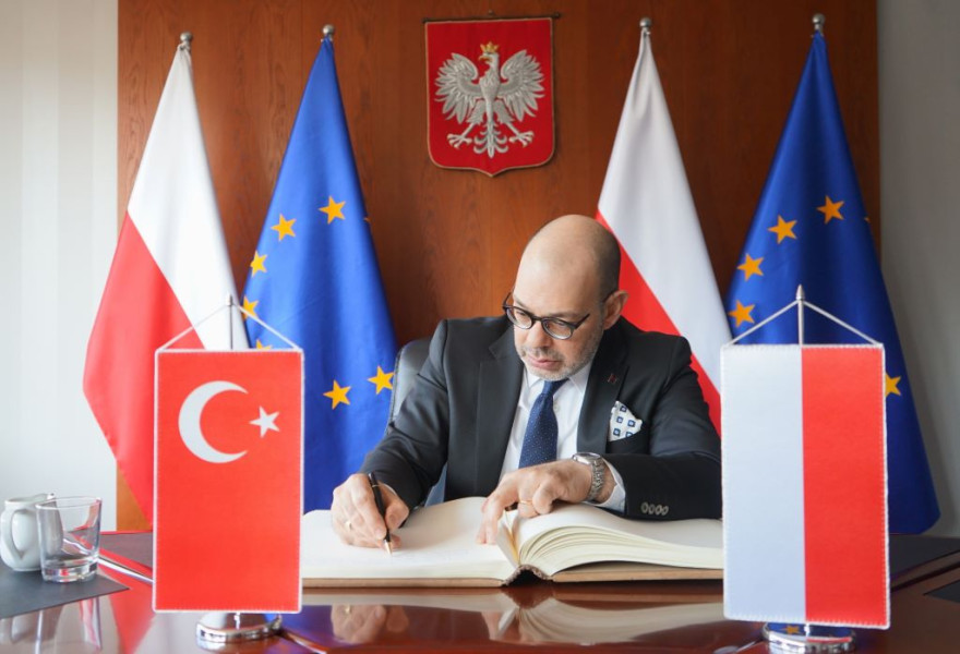 Ambasador Turcji wpisuje się do księgi pamiątkowej, w tle flagi Polski, UE i Godło Polski
