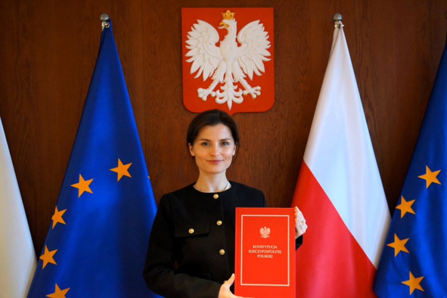 Wojewoda wielkopolska Agata Sobczyk trzyma w rękach przemiot akcji - konstytucję RP, w tle flagi Polski i Uni Europejskiej i Godło Polski